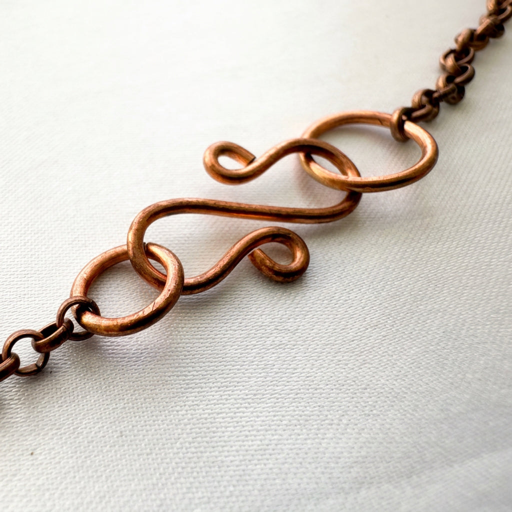 Close-up of a handmade copper S-hook clasp with curled ends, connecting rings and copper chain, resting on pale fabric.