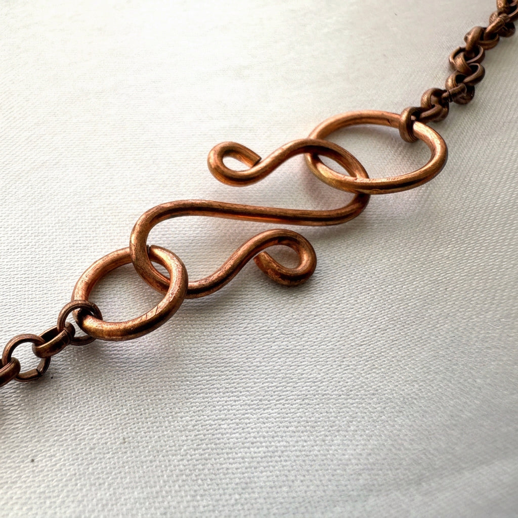 Close-up of a handmade copper S-hook clasp with curled ends, connecting rings and copper chain, resting on pale fabric.