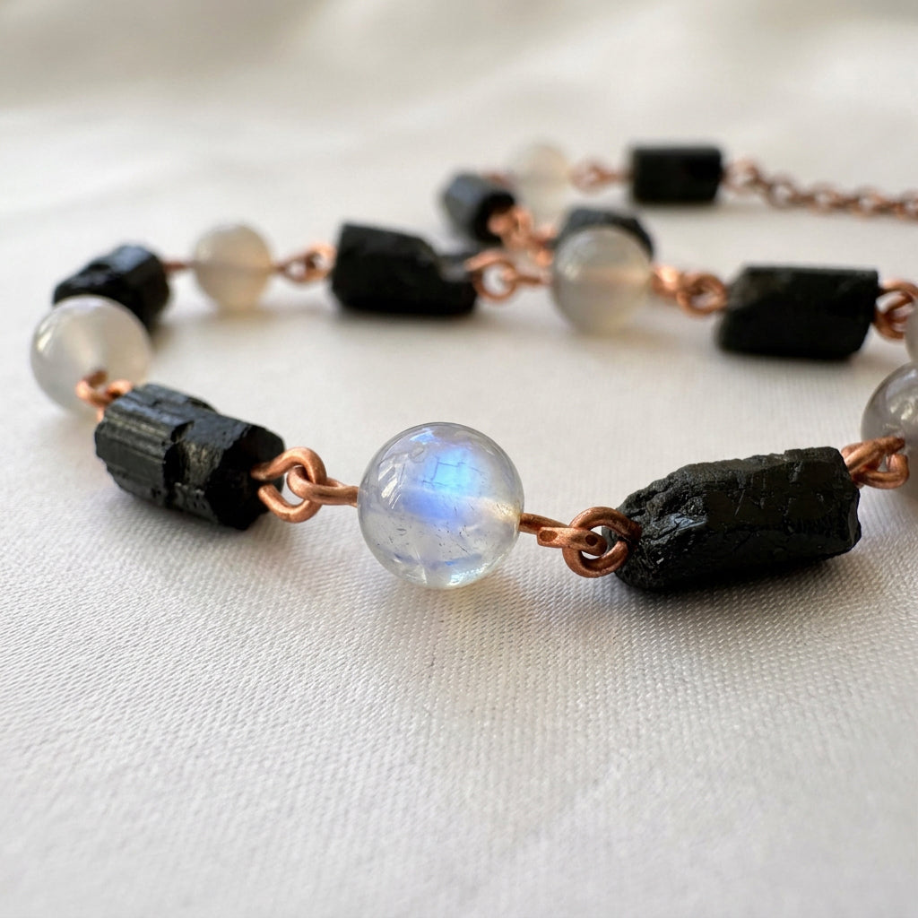 Close-up of a labradorite bead with a vivid blue flash between raw black tourmaline crystals, joined by copper wire links on pale fabric.