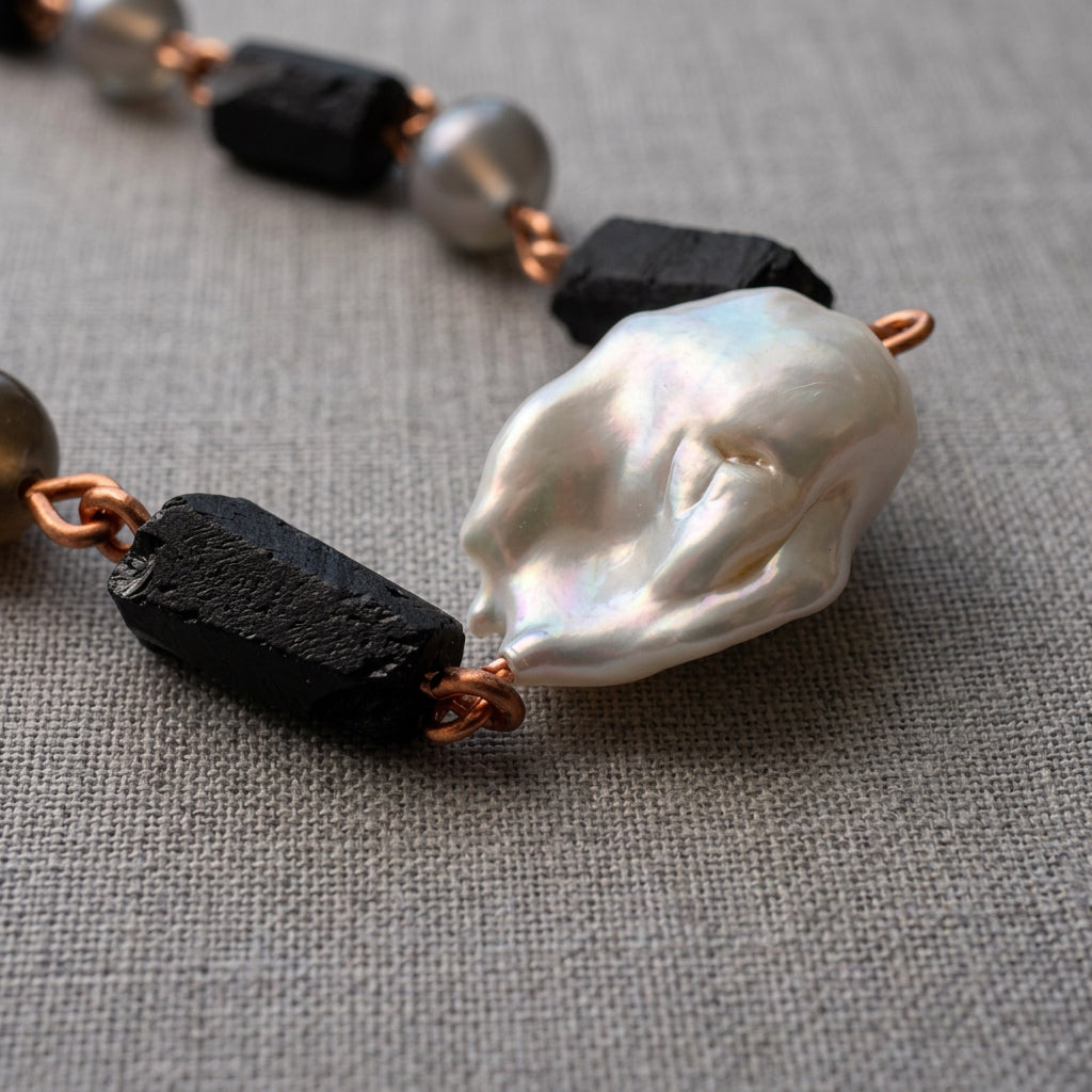 Close-up of a large white Keshi pearl with folded iridescent nacre, flanked by raw black tourmaline crystals and copper wire links on grey linen.