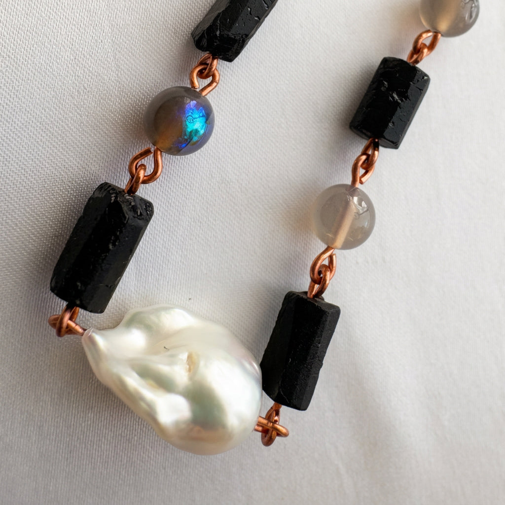Close-up of a white Keshi pearl, raw black tourmaline crystals and grey labradorite beads, one flashing vivid blue-green, joined by copper wire links on pale fabric.