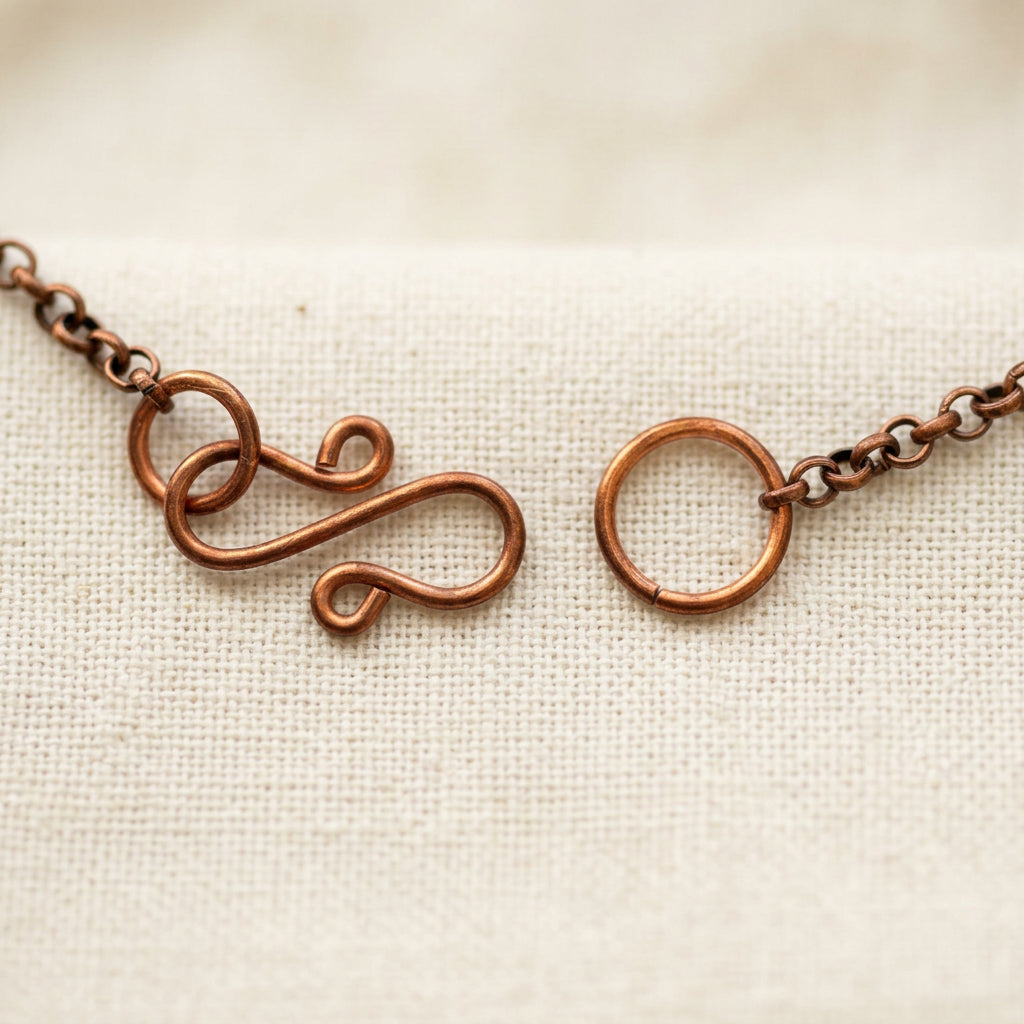 Handmade copper S-hook clasp shown unfastened beside its circular connecting ring, with copper chain extending to either side on cream linen.