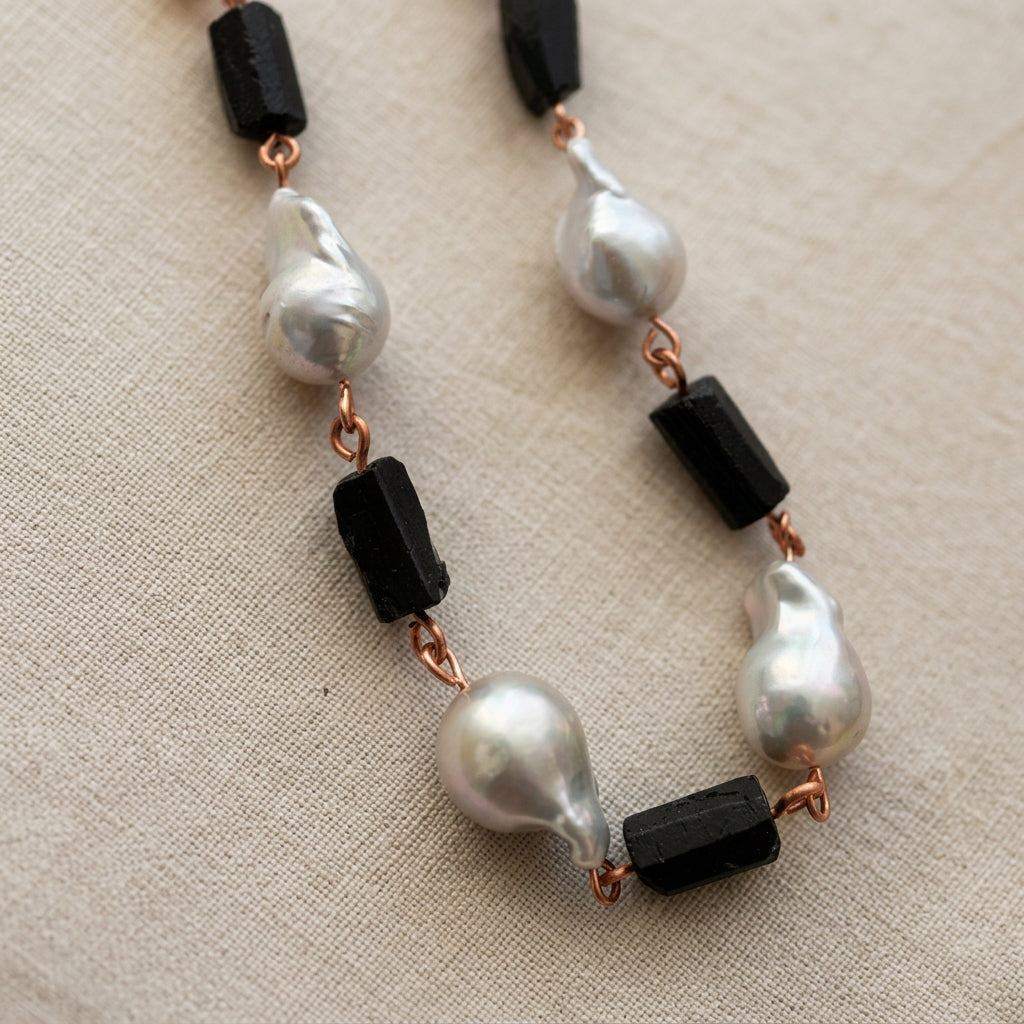 Four large silver Keshi pearls alternating with raw black tourmaline crystals, joined by copper wire links and arranged in a narrow loop on beige linen.