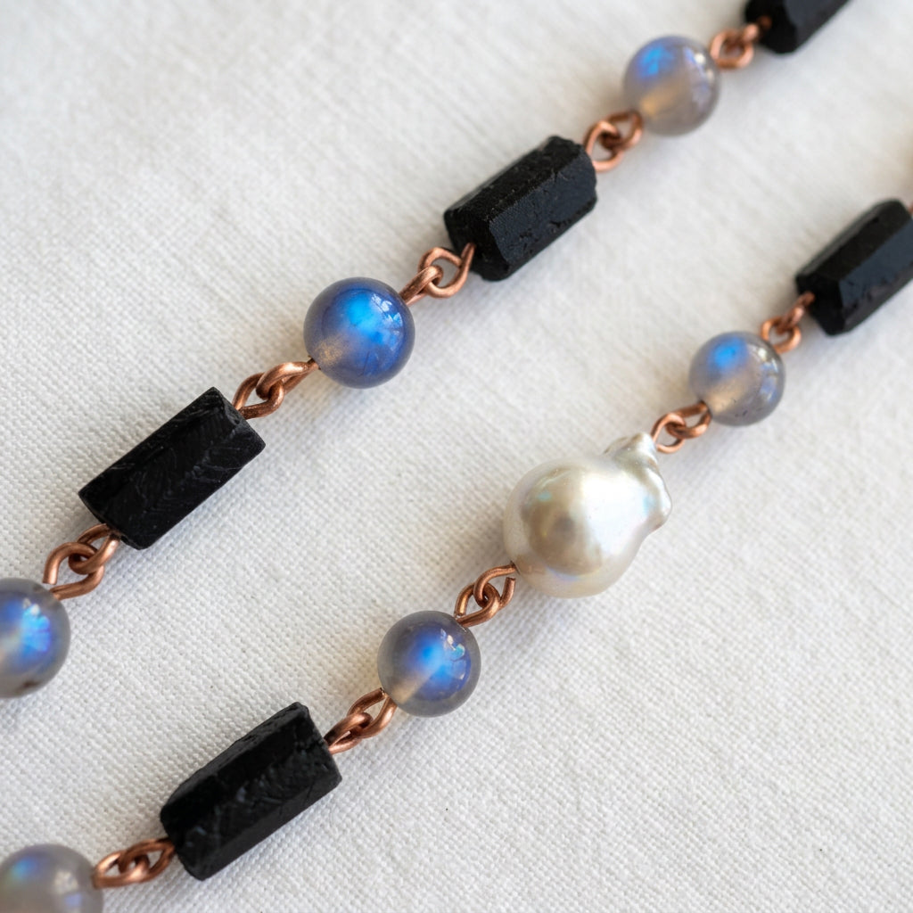 Close-up of raw black tourmaline crystals, labradorite beads flashing vivid blue and a silver Keshi pearl, joined by copper wire links on pale fabric.