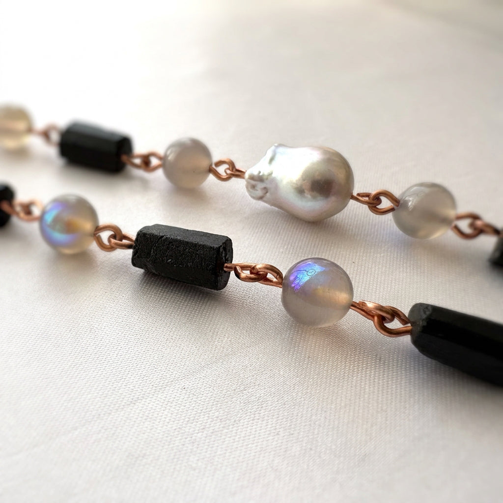 Close-up of raw black tourmaline crystals, grey labradorite beads with blue-purple flashes and a silver Keshi pearl, joined by copper wire links on pale fabric.