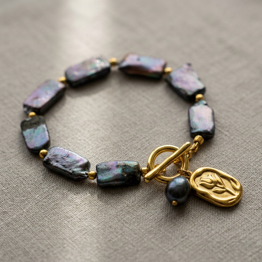 Dark blue rectangular baroque pearl bracelet arranged in a circle on grey linen, with iridescent pearls, gold-coloured spacers and a gold-plated toggle clasp with flower and pearl charms.