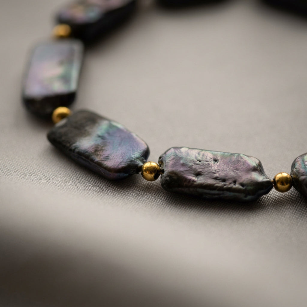 Close-up of dark rectangular baroque pearls with textured surfaces and purple-green iridescence, separated by small gold-coloured beads on grey fabric.