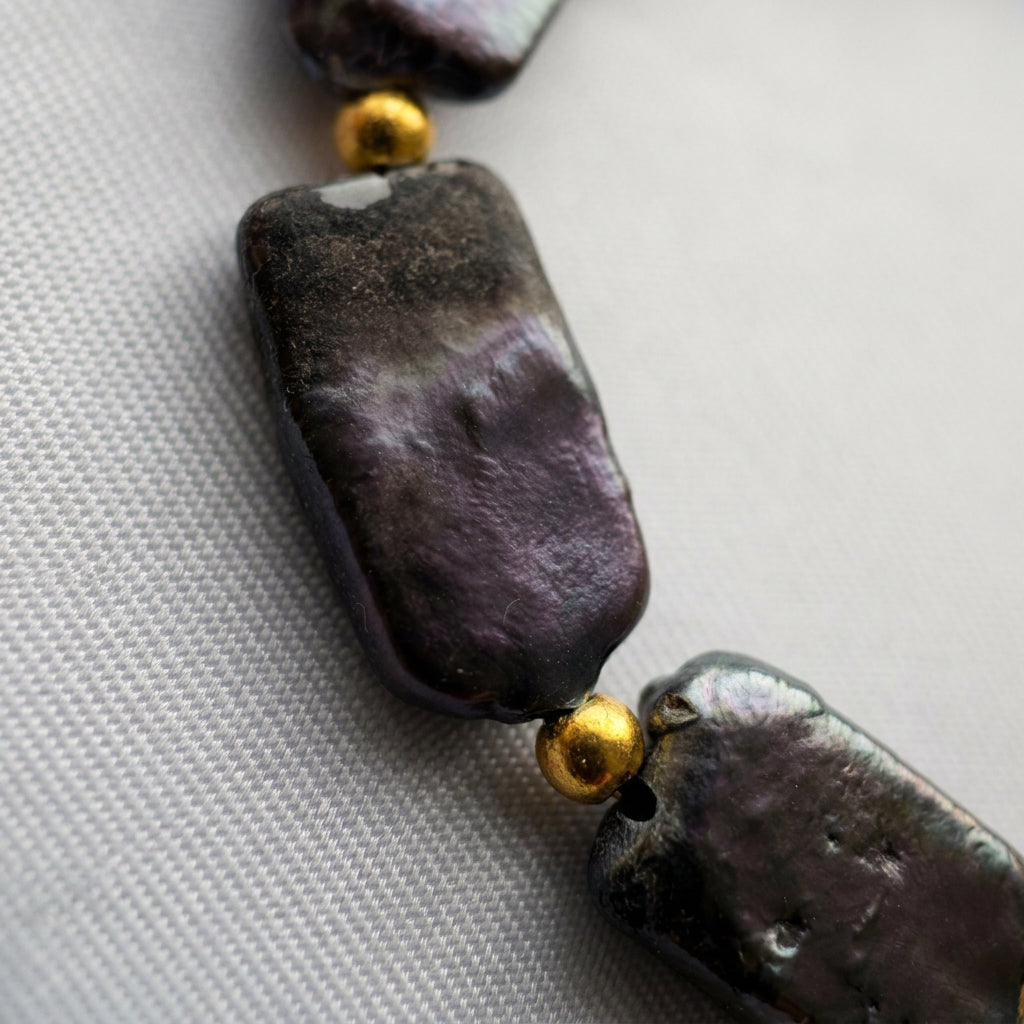 Close-up of dark rectangular baroque pearls with purple iridescence and natural surface dimples, separated by gold-coloured spacer beads on pale fabric.