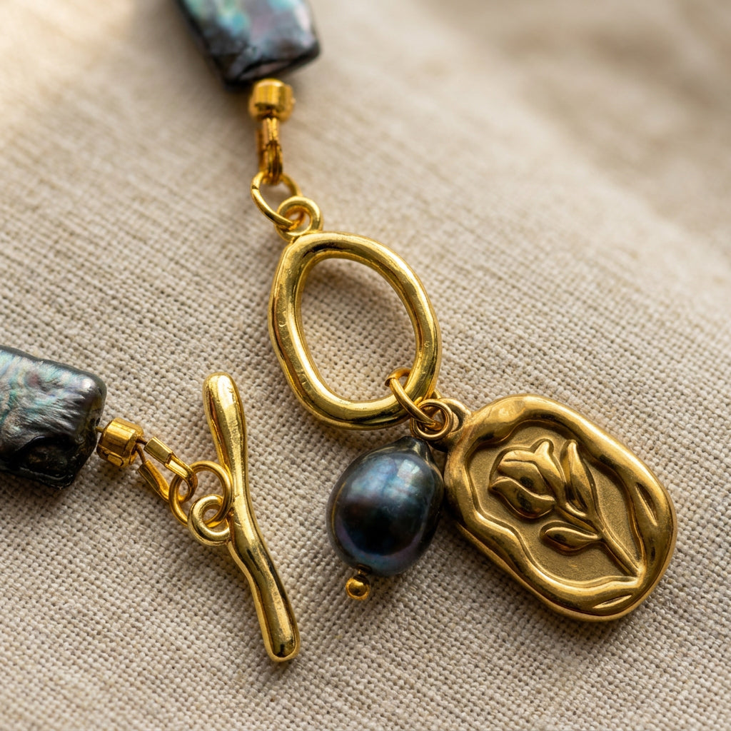 Close-up of an unfastened gold-plated toggle clasp with a raised tulip charm and dangling dark blue pearl, resting on beige linen.
