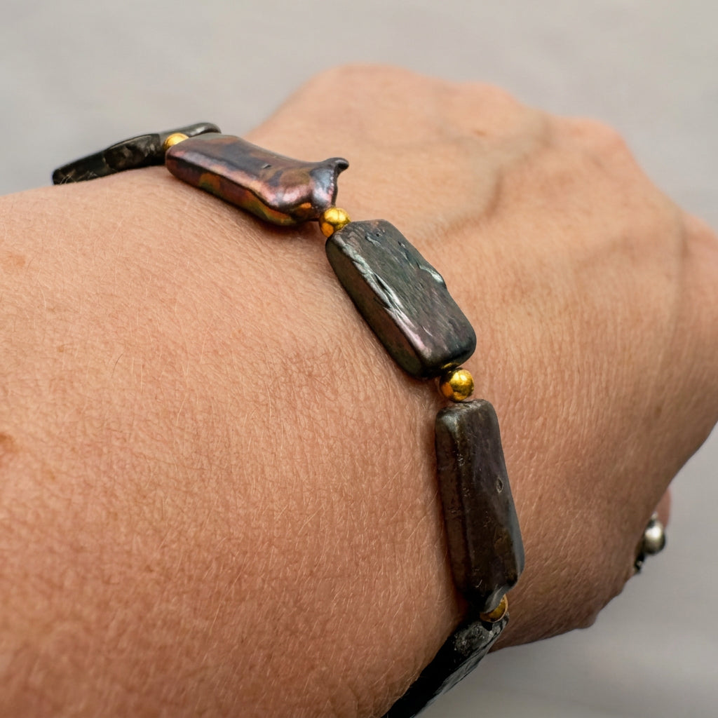 Dark blue baroque pearl bracelet worn on a wrist, showing flat rectangular pearls with green and bronze iridescence, separated by small gold-coloured beads.