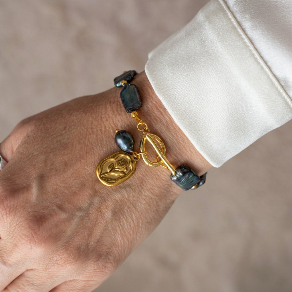Dark blue rectangular baroque pearl bracelet with a fastened gold-coloured toggle clasp, tulip charm and dangling dark pearl, worn beside a white silk shirt cuff.
