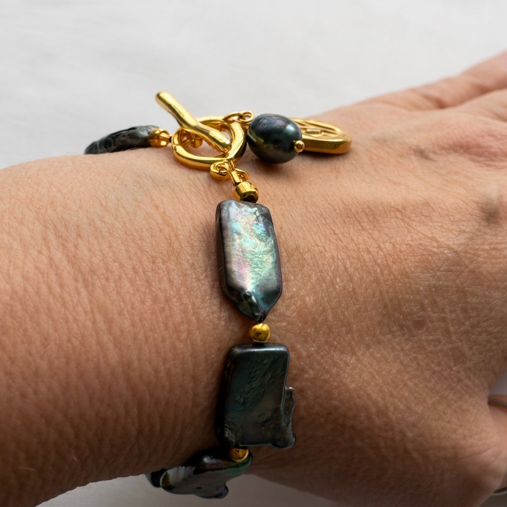 Angled wrist view of a dark blue rectangular baroque pearl bracelet, showing green-pink iridescence, gold-coloured spacer beads and a gold-plated toggle clasp with pearl and flower charms.