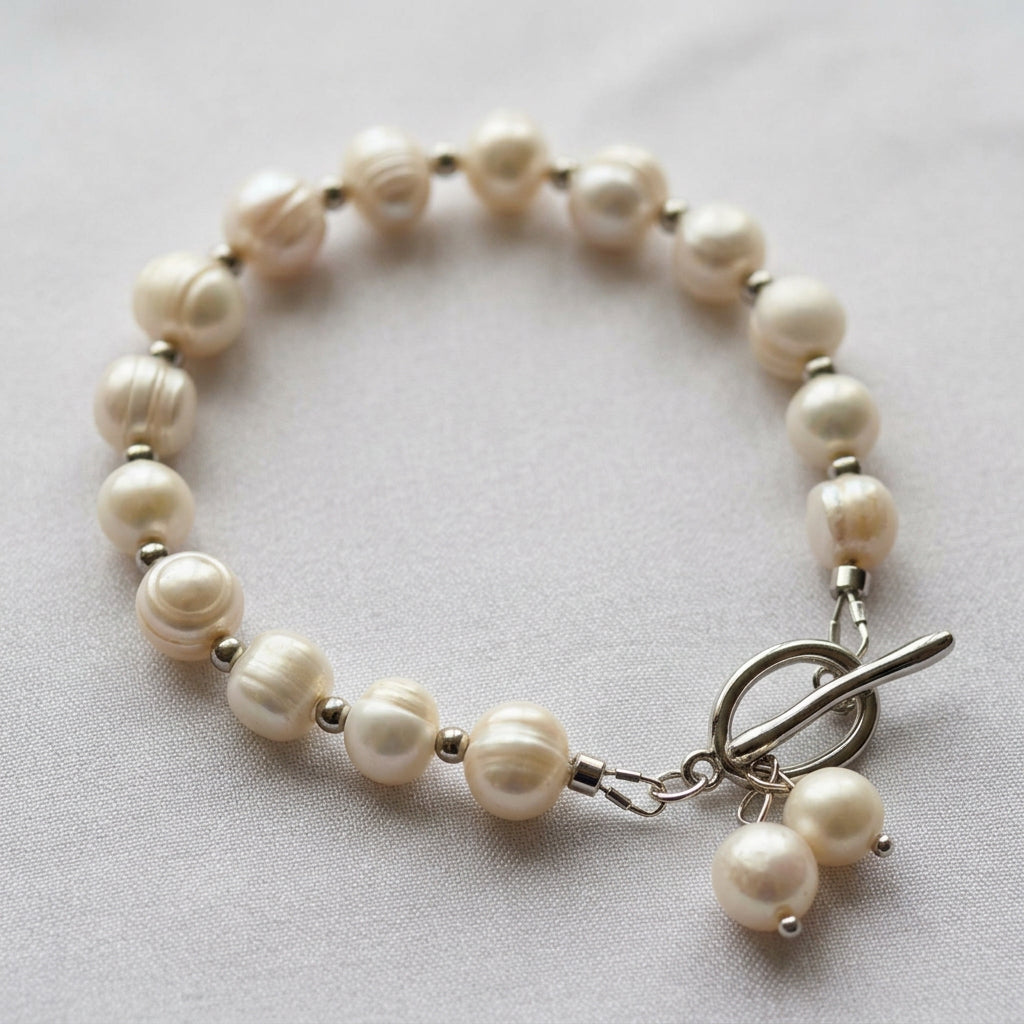 White baroque pearl bracelet arranged in an oval on pale fabric, with silver-coloured spacer beads and a stainless steel toggle clasp finished with two dangling pearl charms.