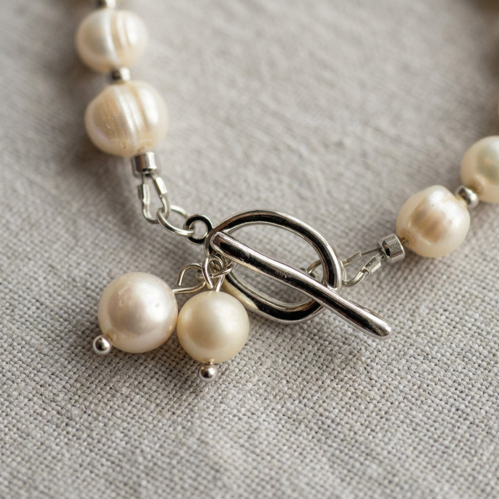 Close-up of a stainless steel circular toggle clasp with two dangling white pearl charms, framed by ridged baroque pearls on grey linen.