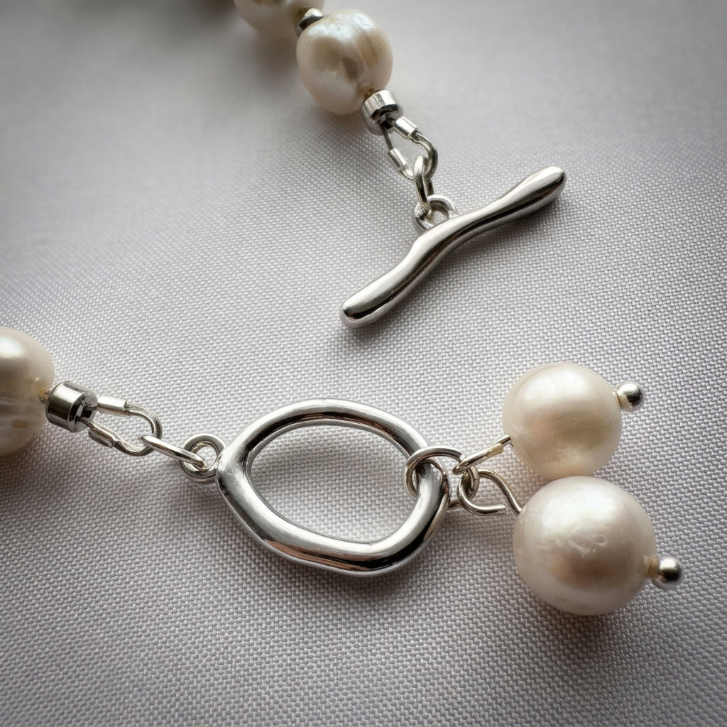Close-up of an unfastened stainless steel toggle clasp, showing the separate curved bar and rounded ring with two dangling white pearl charms on pale fabric.