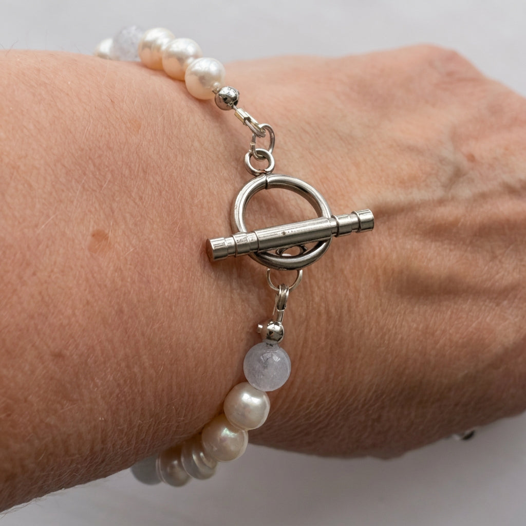 White baroque pearl and aquamarine bracelet worn on a wrist, showing a polished stainless steel circular toggle clasp beside a pale blue aquamarine bead.⠋