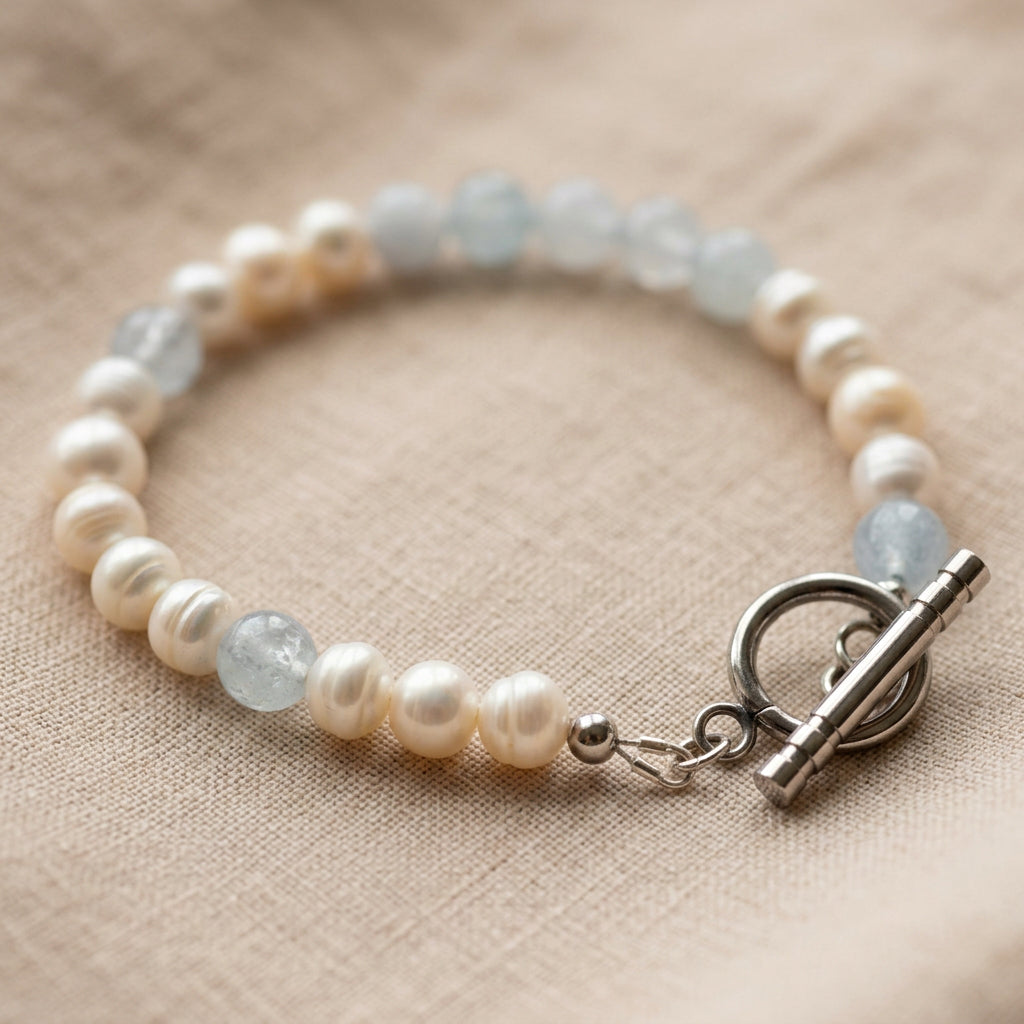 White baroque pearl and pale blue aquamarine bracelet arranged in a circle on beige linen, with a polished stainless steel circular toggle clasp in the foreground.