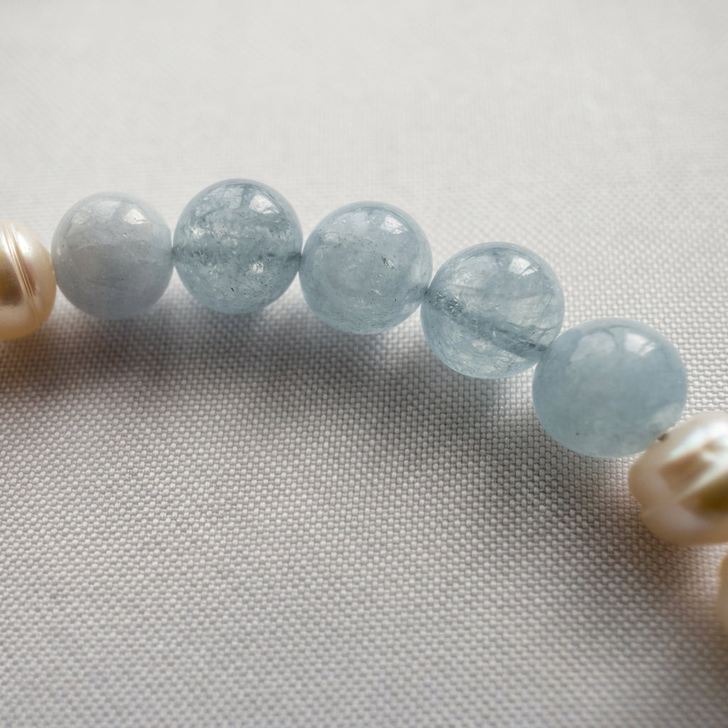 Close-up of five pale blue aquamarine beads with translucent surfaces and visible internal inclusions, between white baroque pearls on pale fabric.⠋