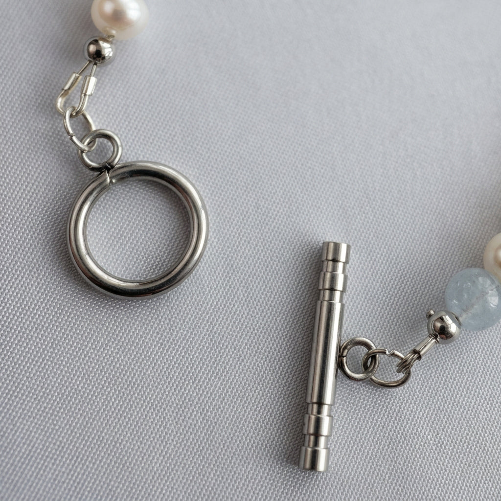 Close-up of an unfastened stainless steel toggle clasp, showing the circular ring and separate bar between white baroque pearls and pale blue aquamarine beads on grey linen.
