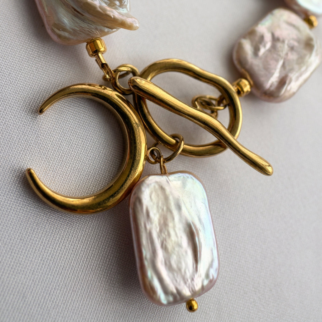 Close-up of a gold-plated toggle clasp with a crescent moon charm and dangling rectangular baroque pearl, showing its ridged, iridescent surface on pale fabric.