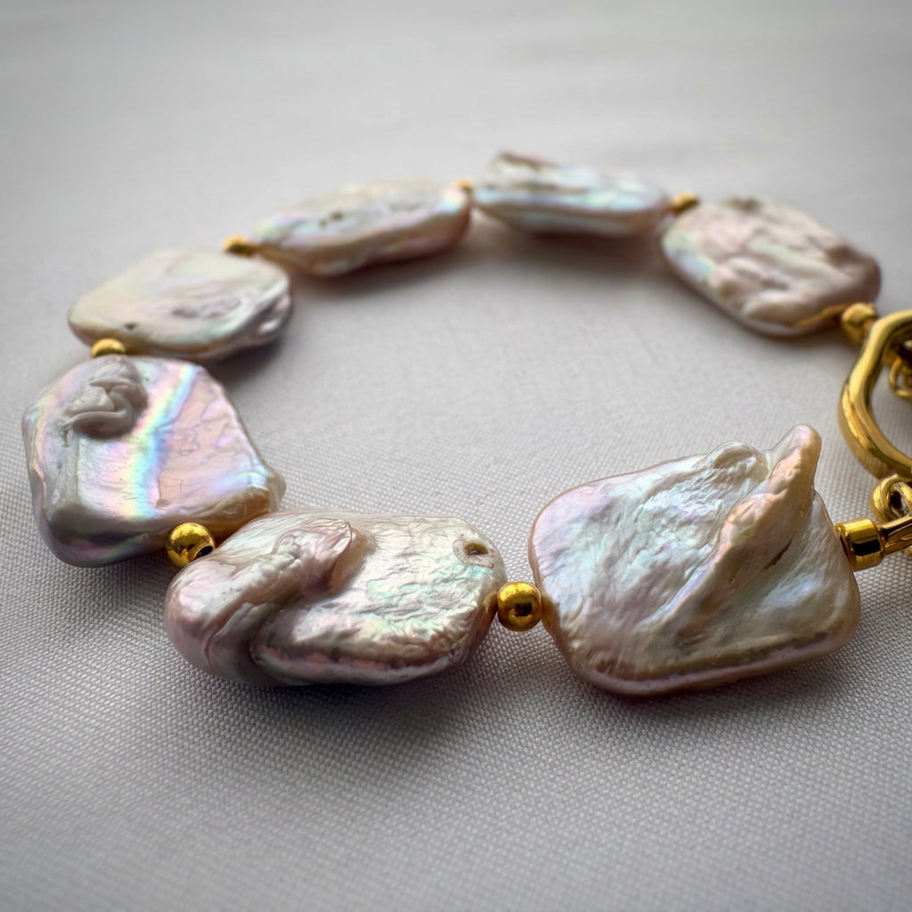 Close-up of rectangular pink baroque pearls arranged in a curve on pale fabric, showing raised natural textures, rainbow iridescence and small gold-coloured spacer beads.