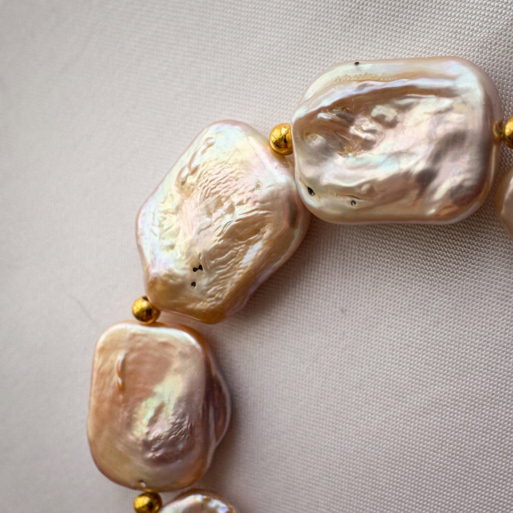 Close-up of three rectangular pink baroque pearls with rippled surfaces and blush-champagne iridescence, separated by small gold-coloured beads on pale fabric.