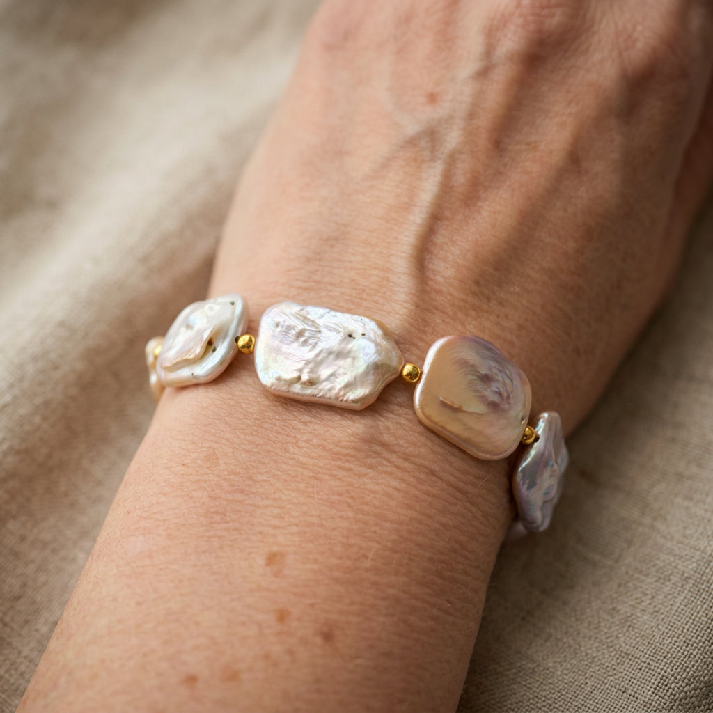 Pink rectangular baroque pearl bracelet worn on a wrist, showing broad, textured pearls with ivory, blush and mauve iridescence, separated by small gold-coloured beads.