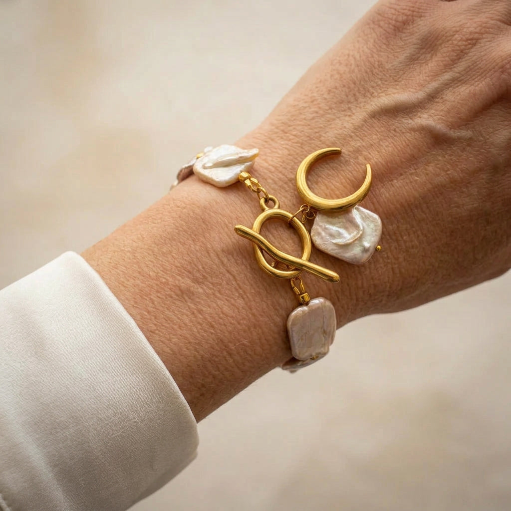 Blush-pink baroque pearl bracelet with a fastened gold-coloured toggle clasp, crescent moon and large white pearl charm, worn on the wrist beside a white silk shirt cuff.