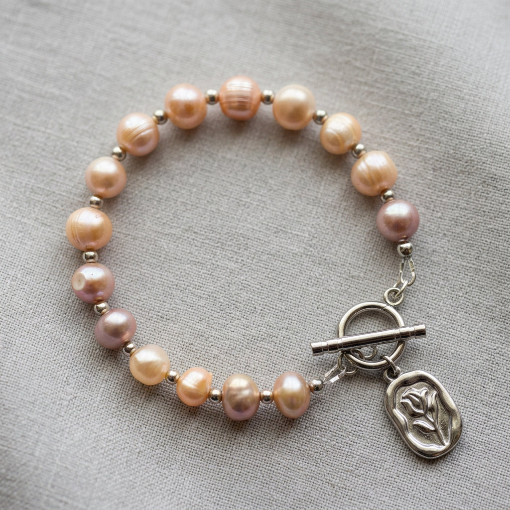 Pink baroque pearl bracelet arranged in a circle on grey linen, with silver-coloured spacer beads, a stainless steel toggle clasp and a rectangular flower pendant.