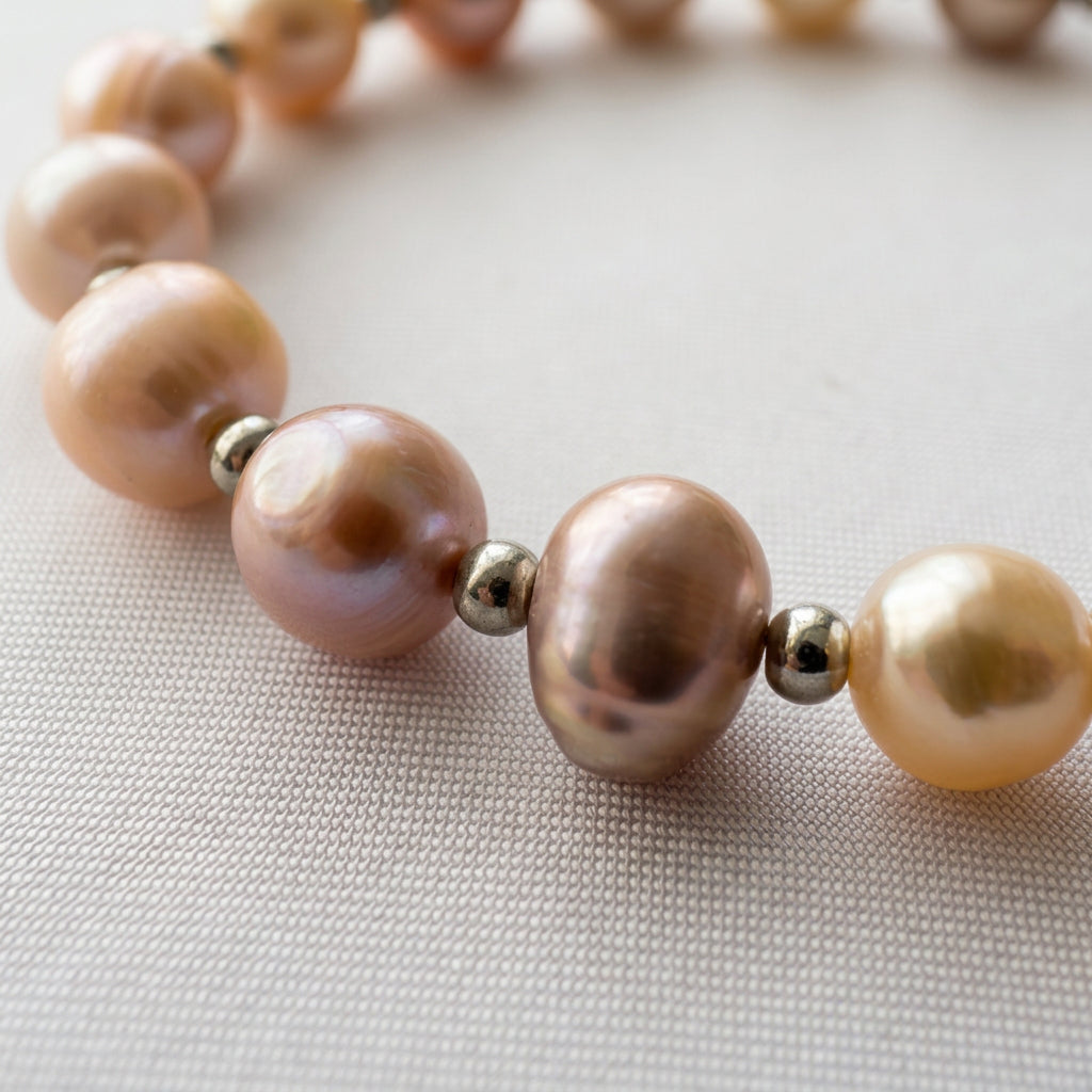 Close-up of lustrous pink baroque pearls in blush, mauve and peach tones, separated by polished silver-coloured beads on pale fabric.