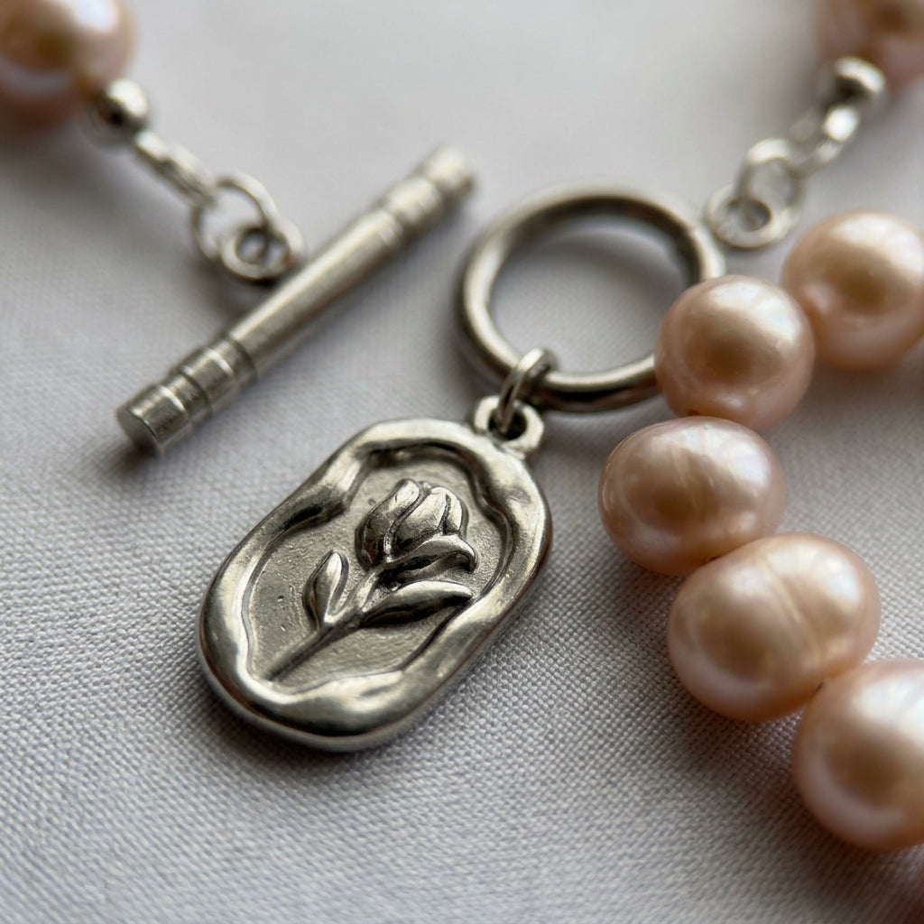Close-up of an unfastened stainless steel toggle clasp and rectangular tulip pendant beside pink baroque pearls on pale fabric.