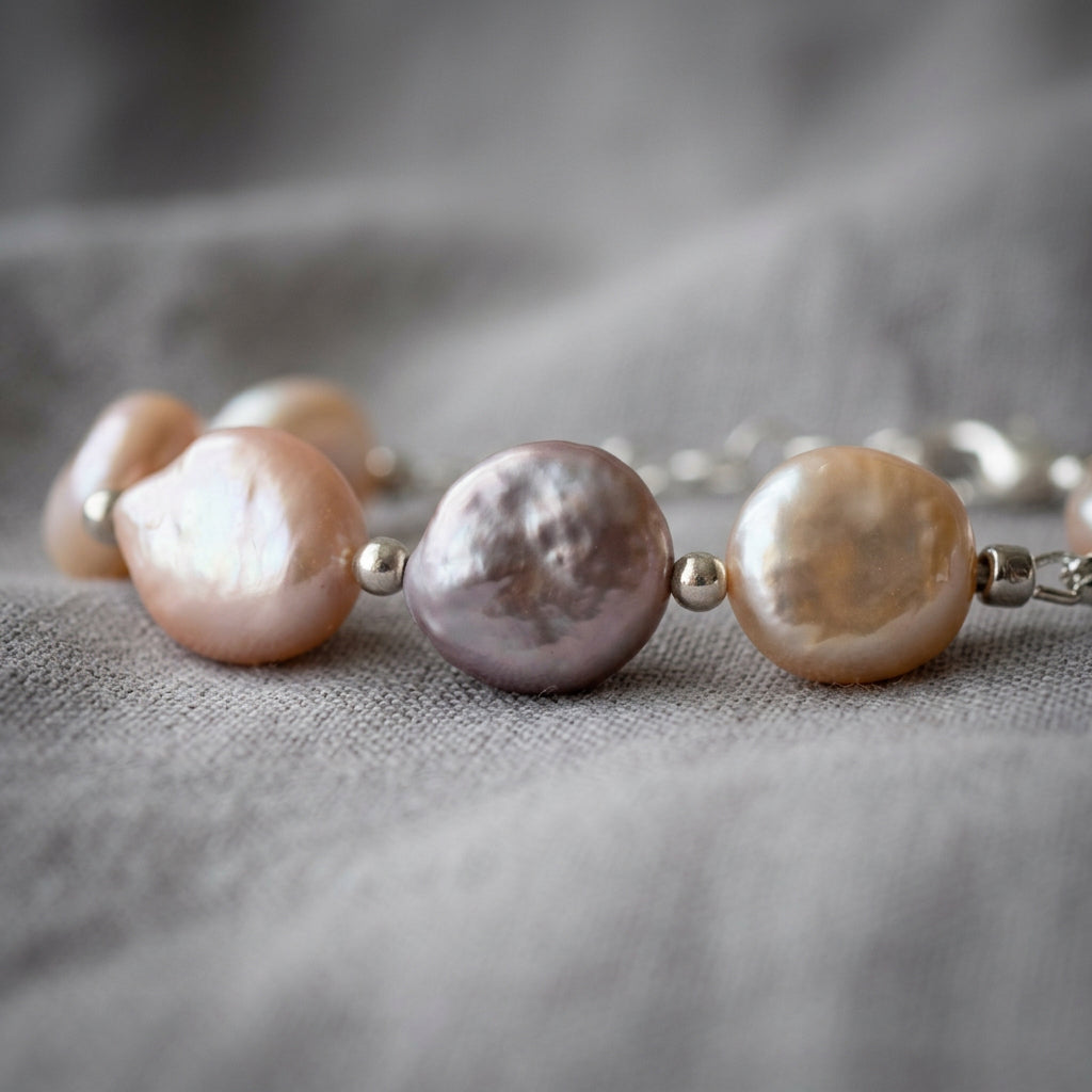 Close-up of pink coin pearls in blush, mauve and peach tones, showing textured iridescent faces separated by silver-coloured beads on grey linen.