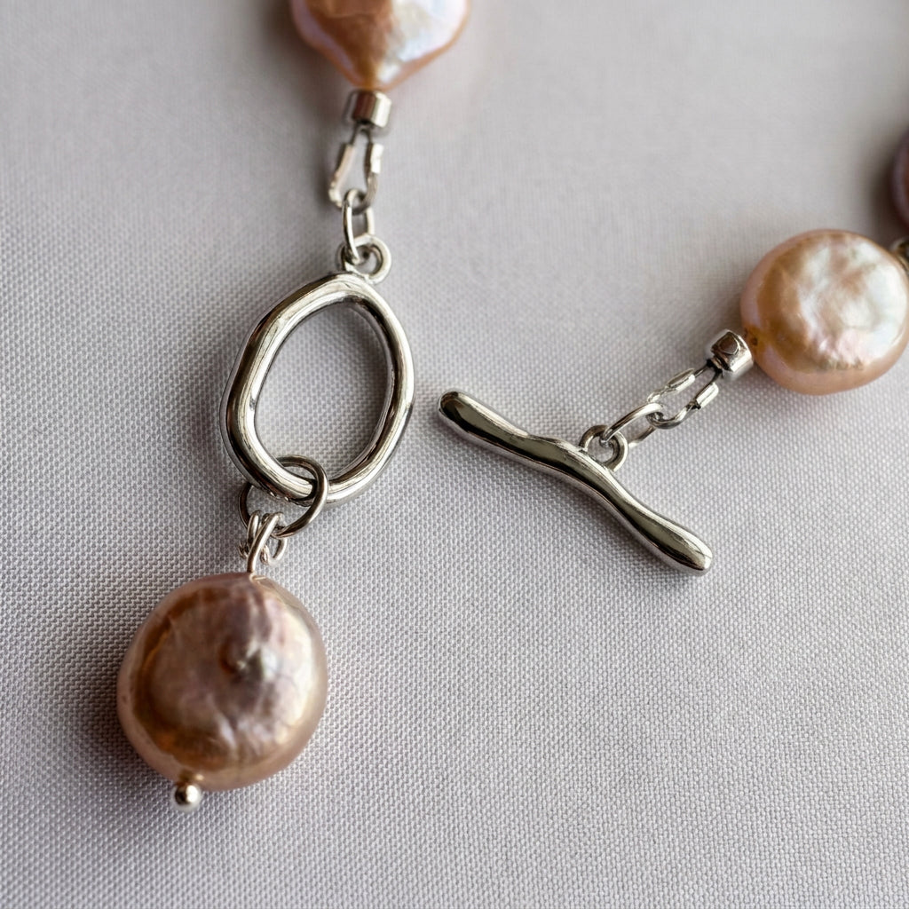 Close-up of an unfastened stainless steel toggle clasp with an oval ring and dangling pink coin pearl charm, resting on pale fabric.