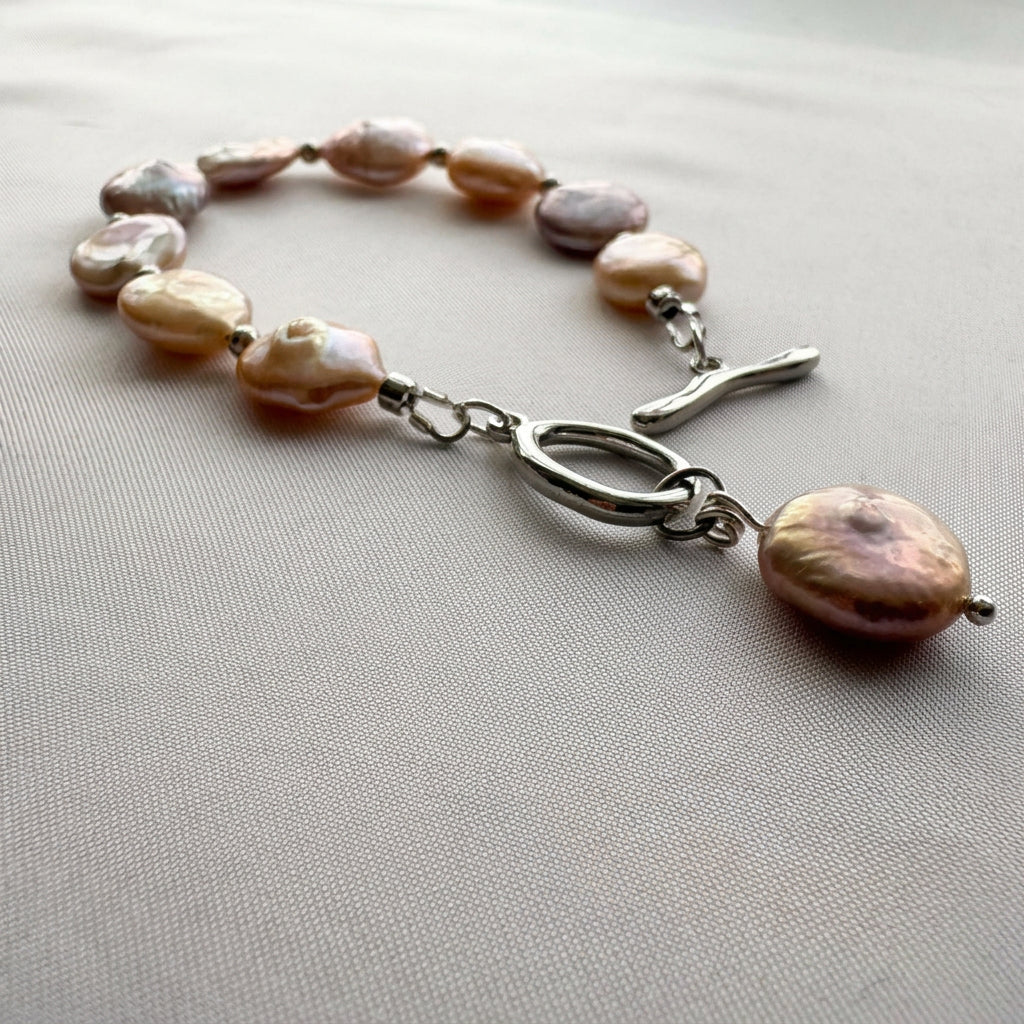 Pink coin pearl bracelet laid open on pale fabric, showing varied blush and mauve pearls, silver-coloured spacers and a stainless steel toggle clasp with a dangling coin pearl charm.