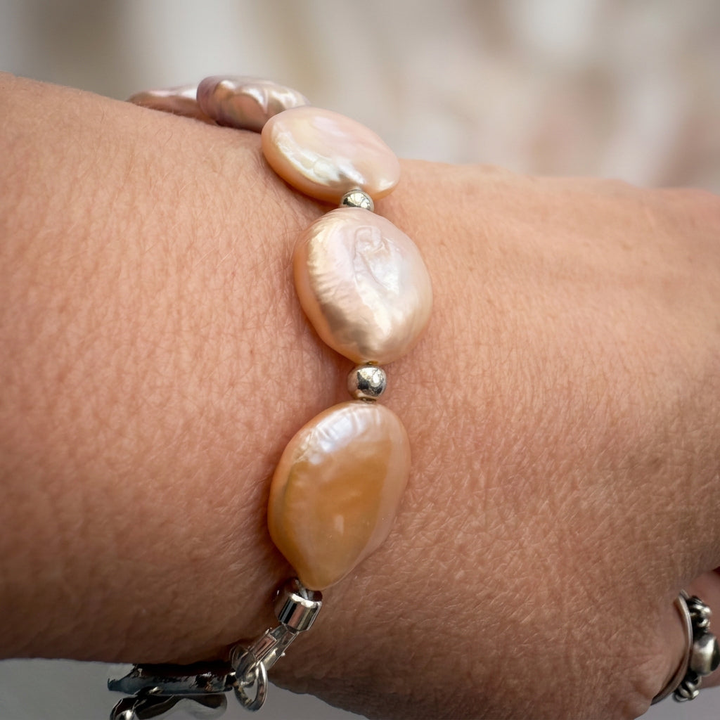 Close-up of a pink coin pearl bracelet worn on a wrist, showing flat blush and peach pearls with soft iridescence, separated by polished silver-coloured beads.