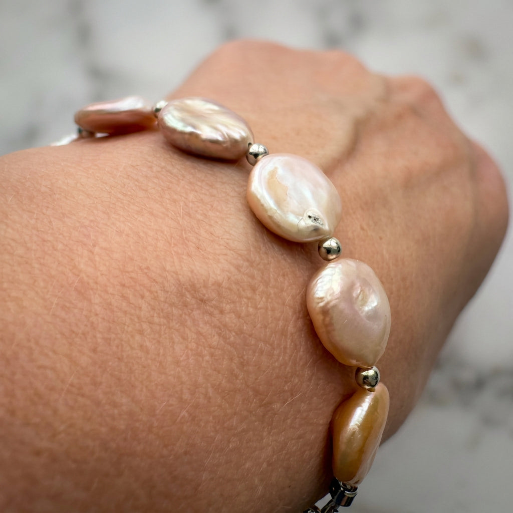 Angled close-up of a pink coin pearl bracelet worn on a wrist, showing flat pearls with rippled cream-pink iridescence and polished silver-coloured spacer beads.