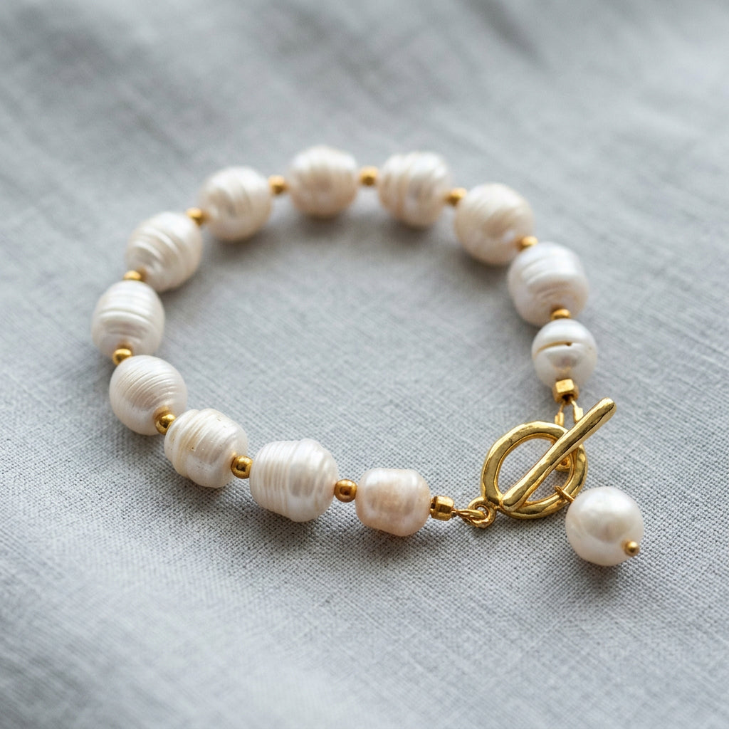 White baroque pearl bracelet arranged in a circle on grey linen, with gold-coloured spacer beads, a gold-plated toggle clasp and a dangling white pearl charm.