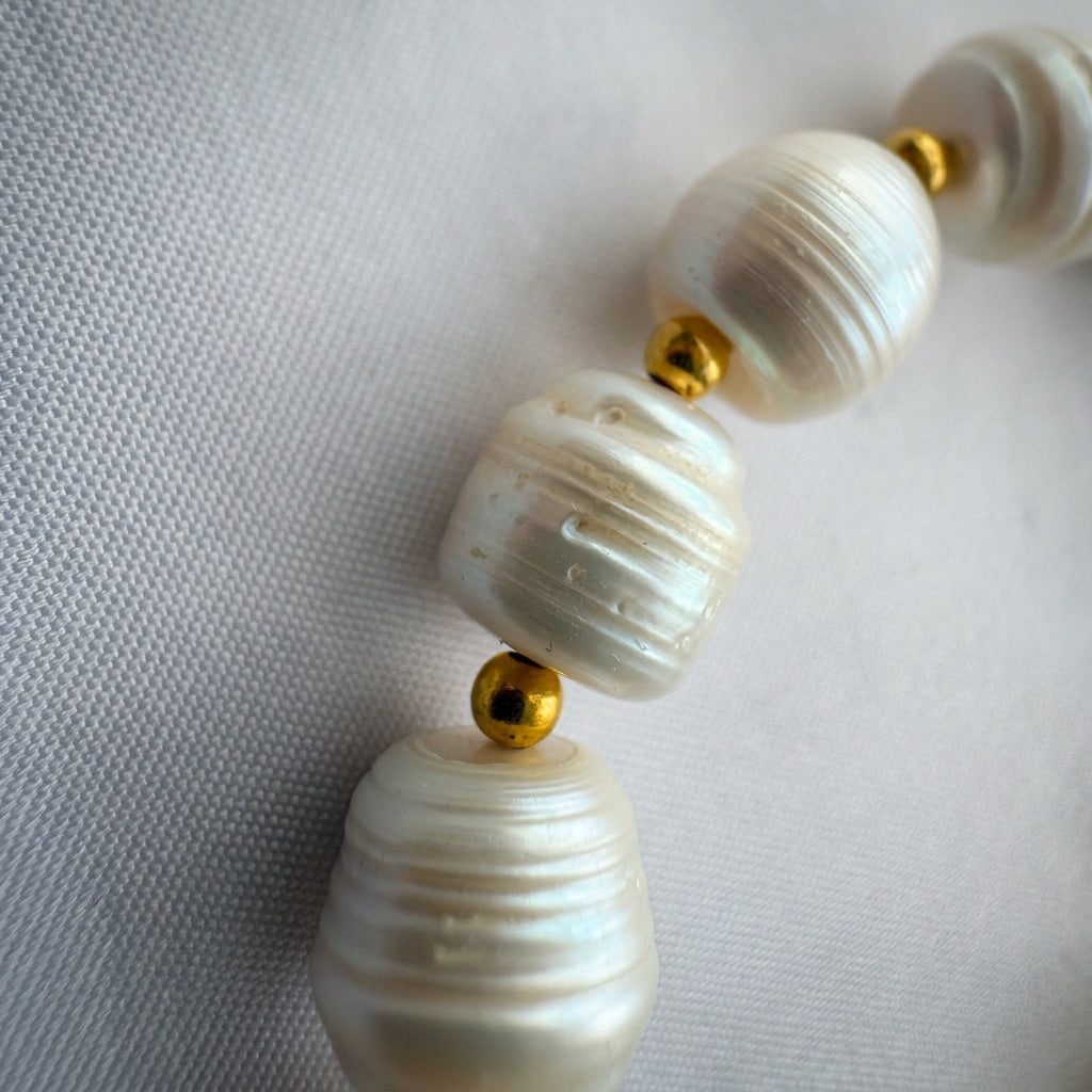 Close-up of white baroque pearls with natural ridges, irregular shapes and soft iridescence, separated by small gold-coloured beads on pale fabric.