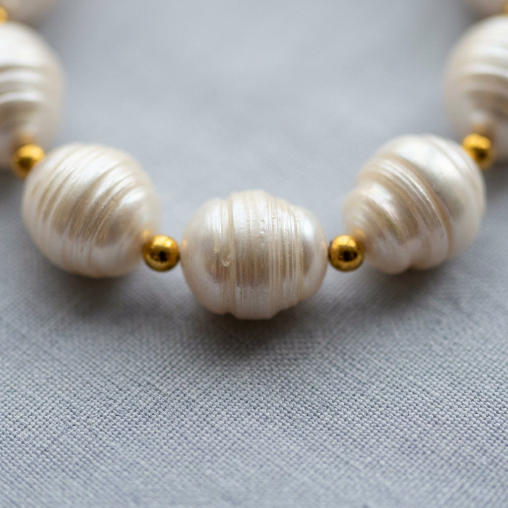 Close-up of three white baroque pearls with pronounced natural ridges and soft lustre, separated by small gold-coloured beads on grey linen.