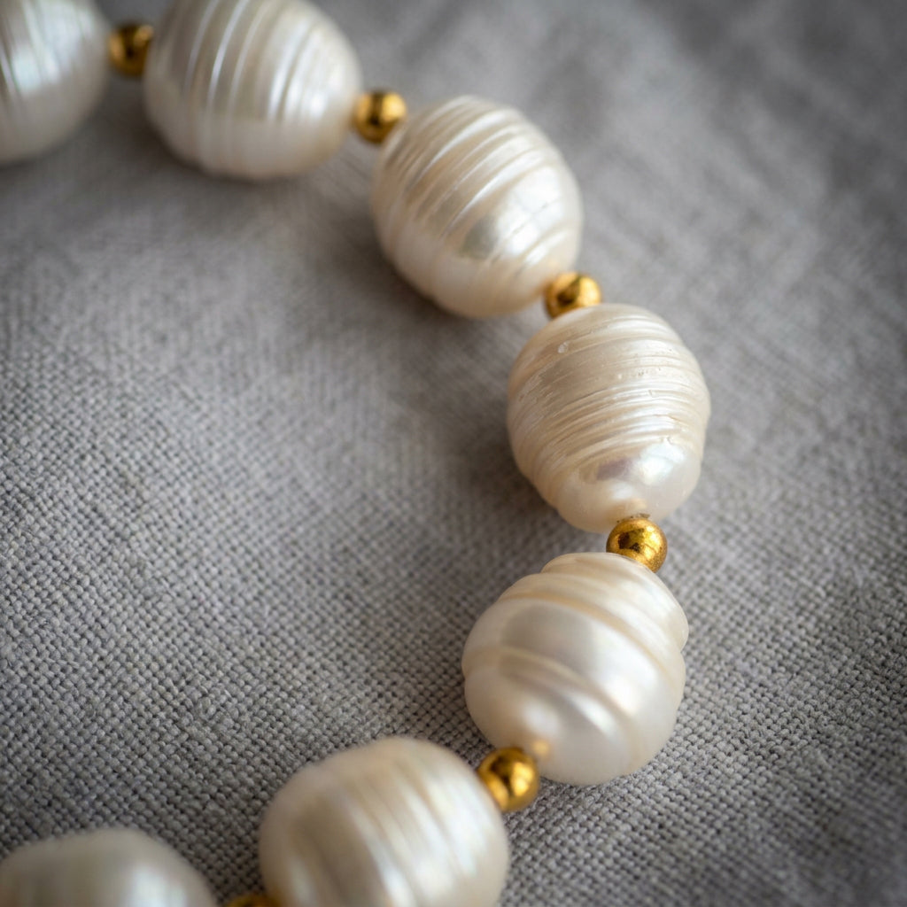 White baroque pearls arranged in a curve on grey linen, showing varied oval shapes, natural ridges and soft lustre between small gold-coloured spacer beads.
