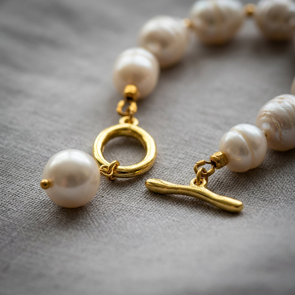 Close-up of an unfastened gold-plated toggle clasp with a dangling white pearl charm, attached to a white baroque pearl bracelet on grey linen.