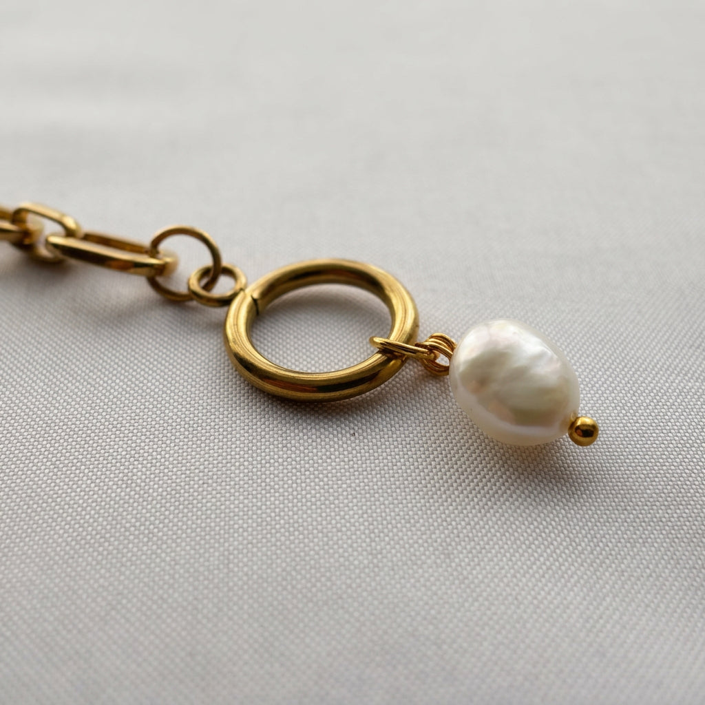 Close-up of the gold-plated toggle clasp ring with a dangling white baroque pearl charm, attached to a paperclip chain on pale fabric.