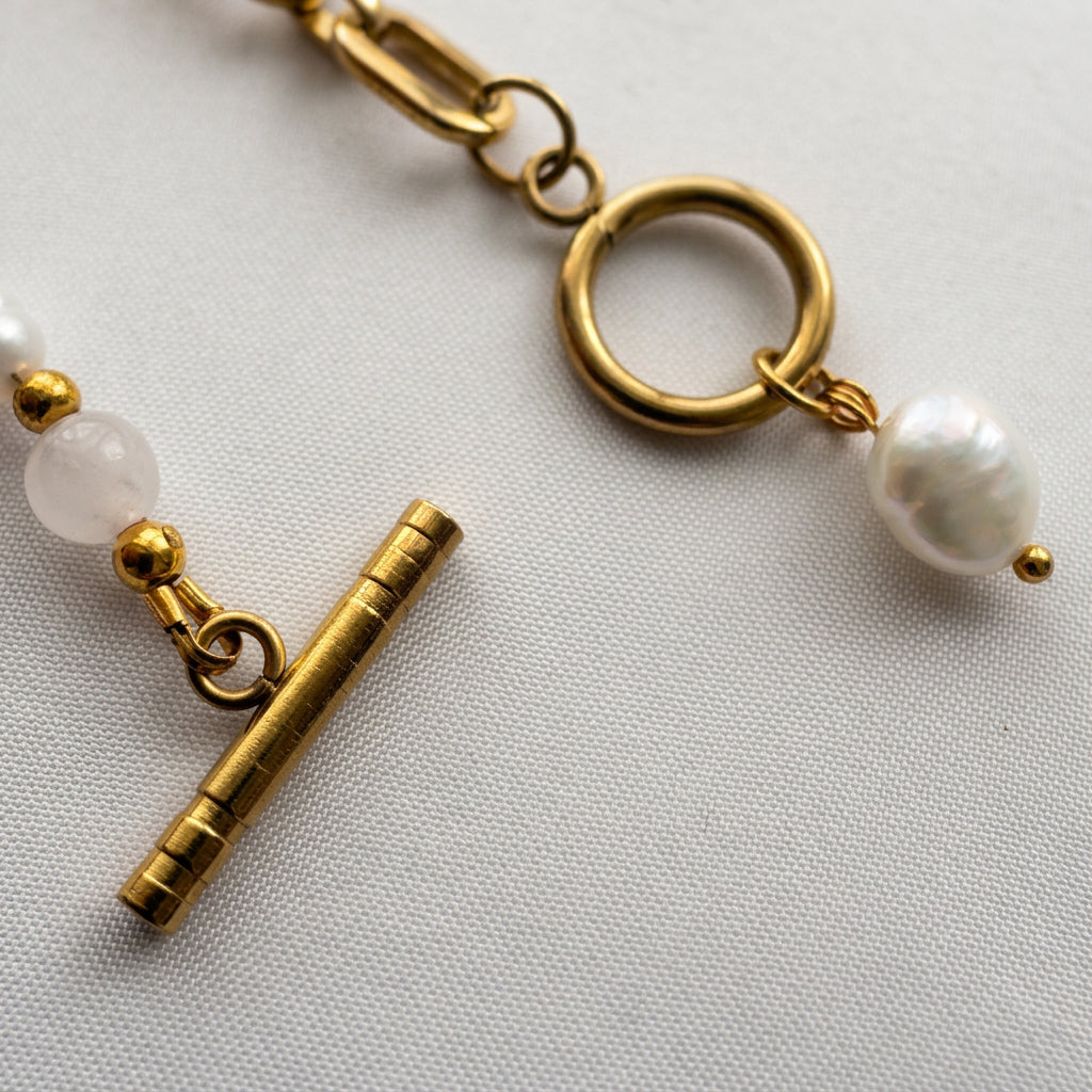 Close-up of an unfastened gold-plated toggle clasp on pale fabric, with pink quartz beside the bar and a white baroque pearl charm hanging from the ring.