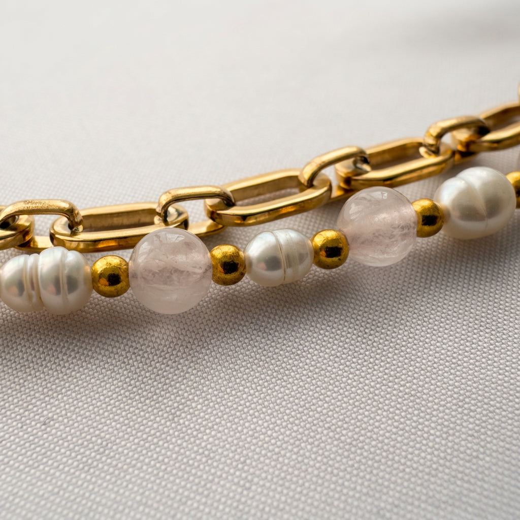 Close-up of pale pink quartz beads and white baroque pearls separated by gold-coloured beads, laid alongside the bracelet’s gold-plated paperclip chain on pale fabric.