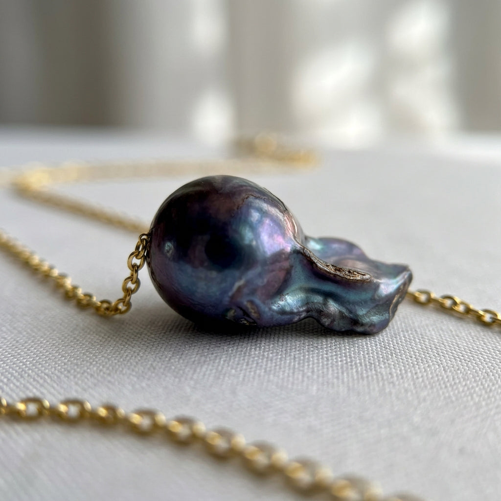 Close-up of a dark blue keshi pearl on a gold-filled chain, showing its rounded dome and folded nacre wing with teal, violet and pink iridescence on white fabric.