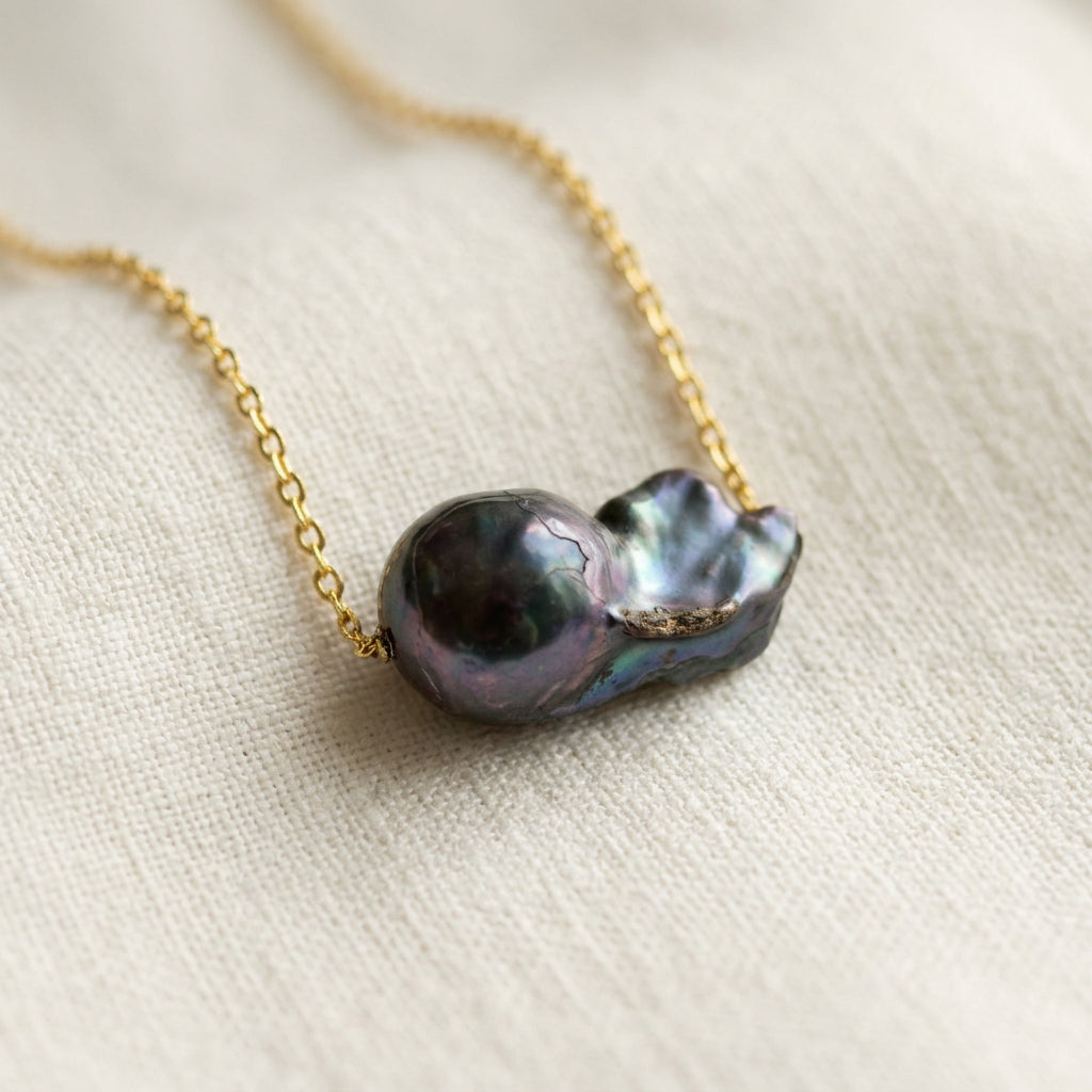 Dark blue keshi pearl on a fine gold-filled chain resting on cream linen, with a glossy rounded dome and curled nacre folds reflecting teal, violet and silver.
