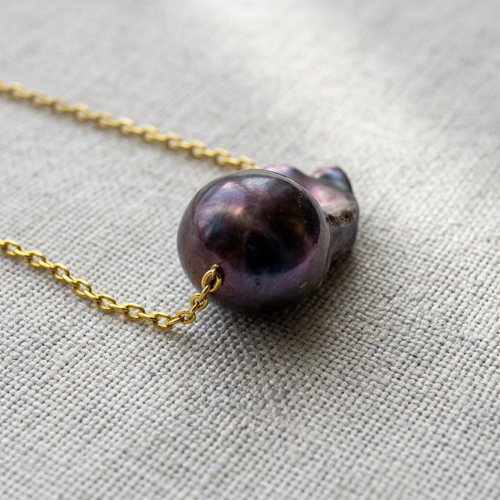 Close-up of the glossy rounded dome of a dark blue keshi pearl on a gold-filled chain, showing deep violet and aubergine reflections with its folded nacre wing behind it on grey linen.