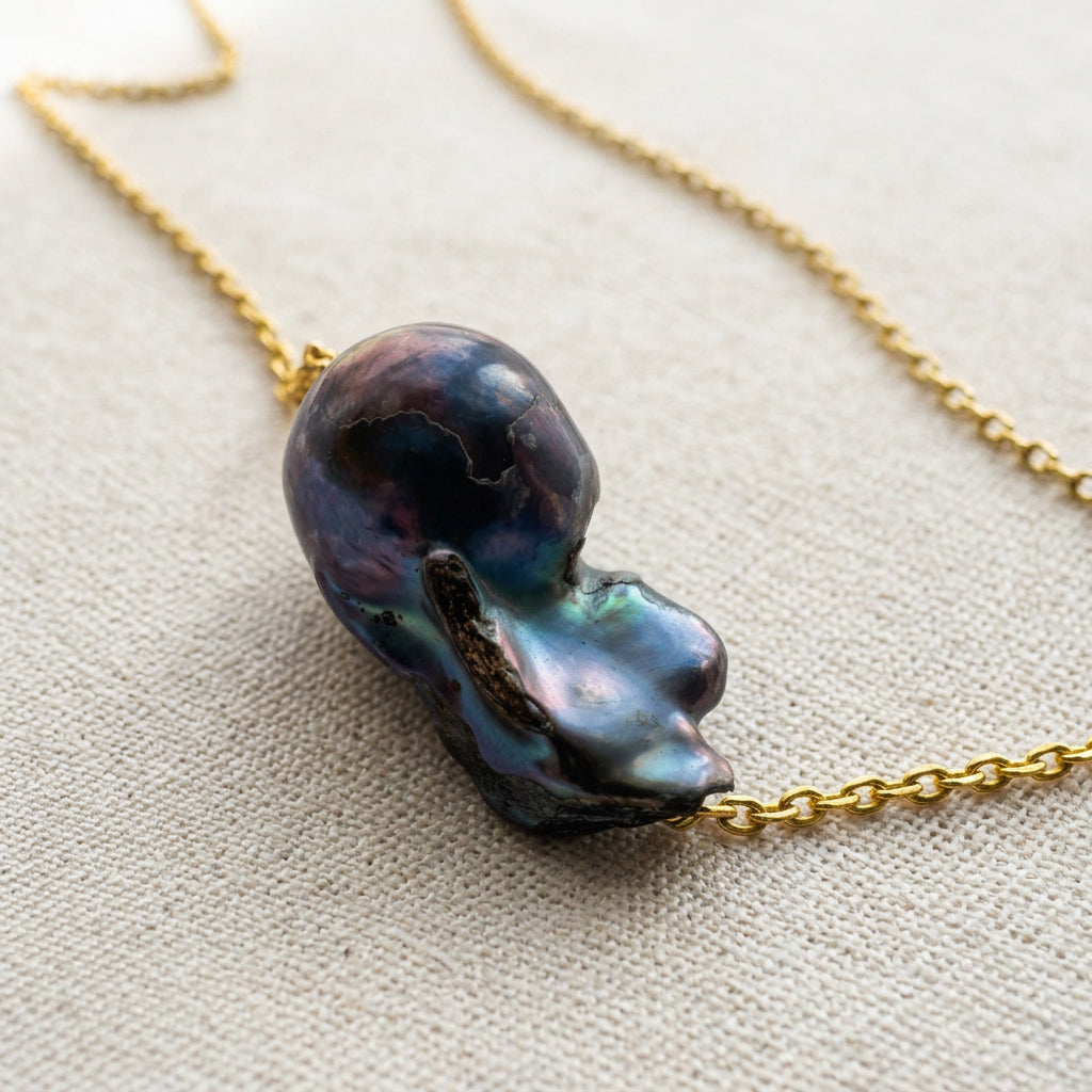 Angled close-up of a dark blue keshi pearl on a gold-filled chain, showing its rounded dome, deep nacre folds and teal, violet and pink reflections on cream linen.