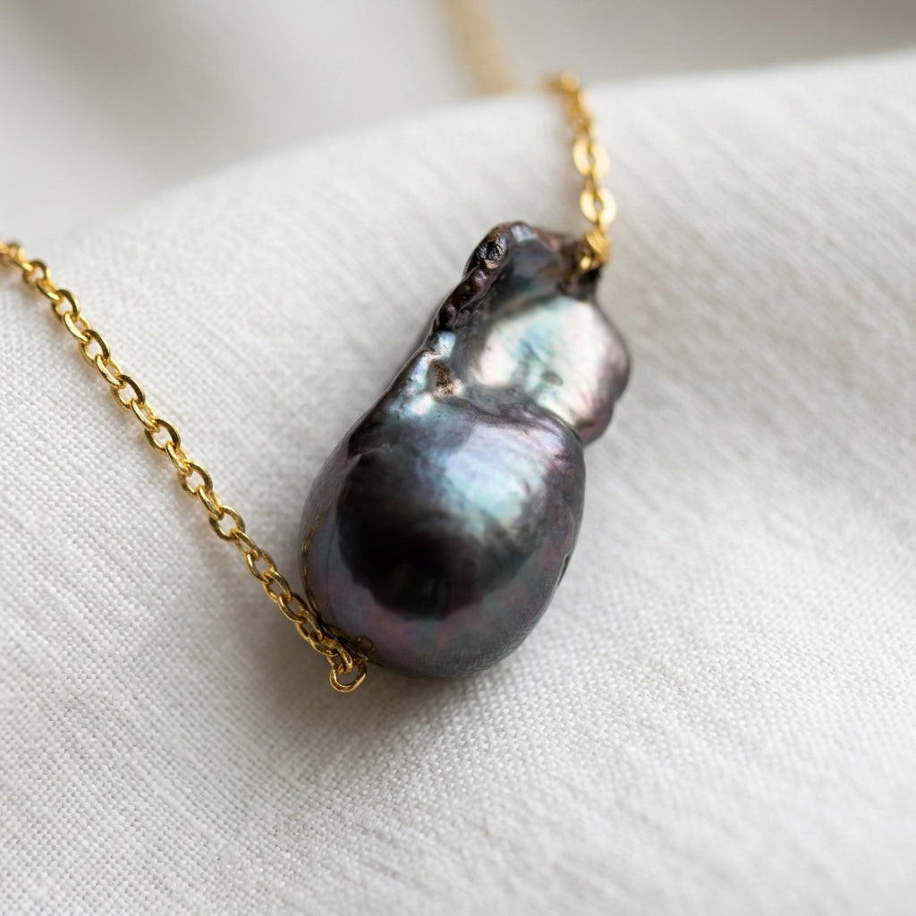 Close-up of a dark blue keshi pearl lying lengthways on white fabric, with a glossy elongated dome, irregular nacre folds and silver-teal reflections, threaded onto a fine gold-filled chain.