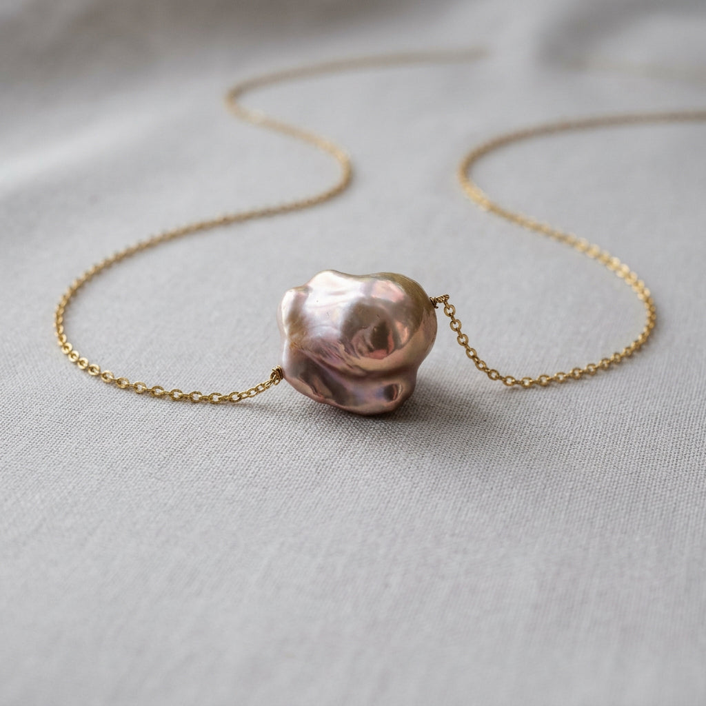 Silvery-blush keshi pearl with rose-gold reflections on a fine gold-over-sterling-silver chain, arranged in loose curves on grey fabric, showing the pearl’s sculptural, irregular shape.